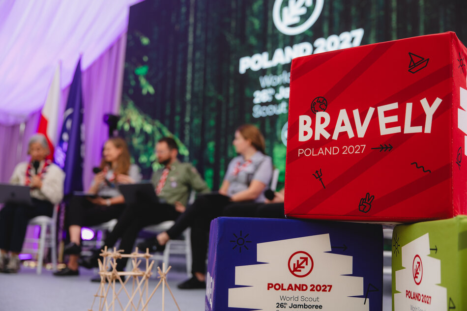 Na scenie siedzą uczestnicy panelu dyskusyjnego w harcerskich mundurach. Na pierwszym planie widoczna jest czerwona kostka z napisem „Bravely Poland 2027”, obok baner z logo Światowego Jamboree Skautowego.