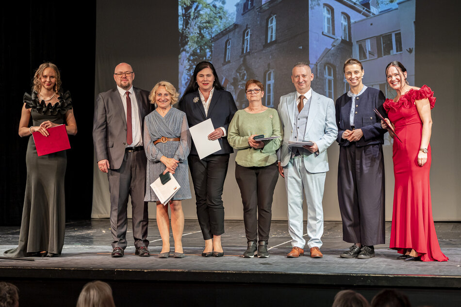 Grupa osób pozuje na scenie podczas uroczystego wydarzenia, trzymając w dłoniach dyplomy i dokumenty. Uczestnicy są elegancko ubrani, a w tle widać projekcję przedstawiającą budynek związany z okazją spotkania. 
