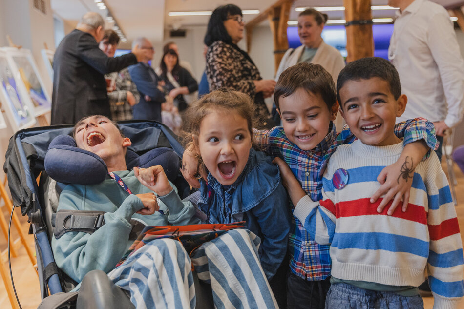Grupa uśmiechniętych dzieci pozuje razem podczas spotkania pełnego radości i pozytywnej energii. W tle widać dorosłych rozmawiających przy wystawie, co nadaje scenie ciepły, towarzyski charakter. 