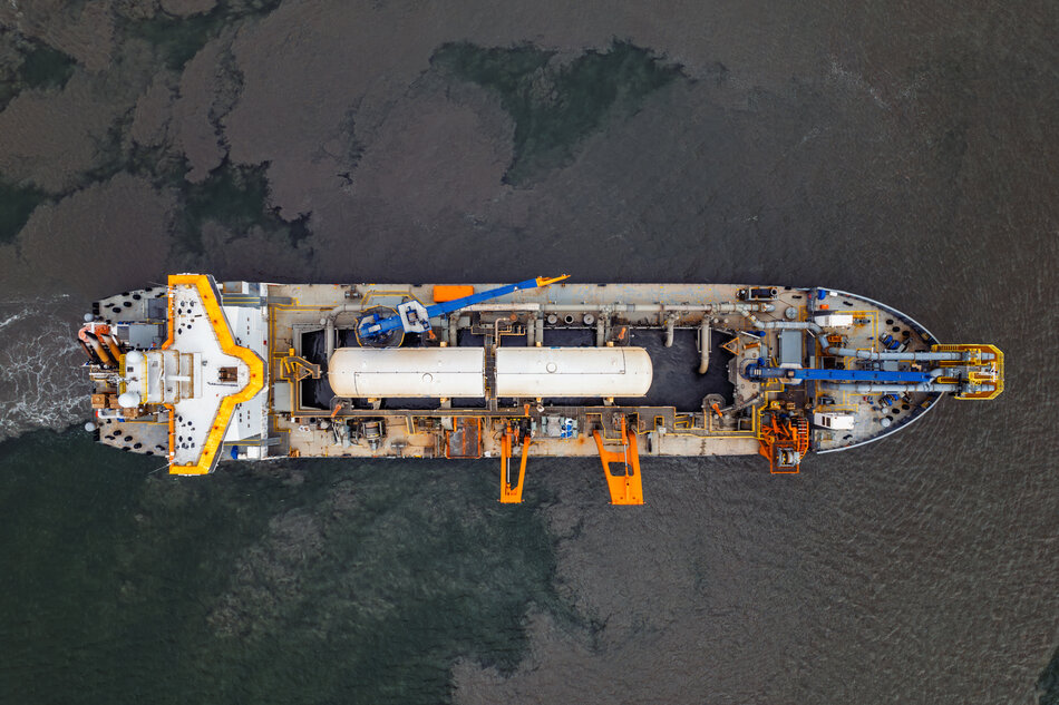 Statek przemysłowy sfotografowany z lotu ptaka płynie po spokojnej wodzie, a jego pokład wypełniają rurociągi, dźwigi i zbiorniki techniczne. Jasne elementy konstrukcji kontrastują z ciemnoszarą powierzchnią morza, podkreślając techniczny charakter jednostki. 