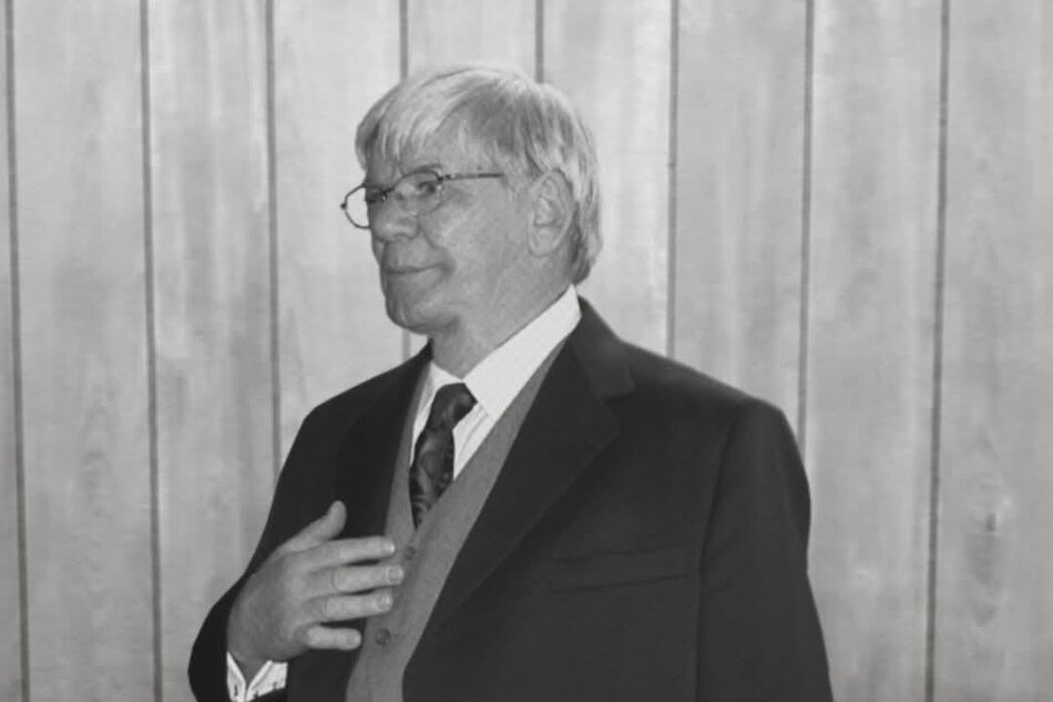 Na zdjęciu widać starszego mężczyznę w garniturze, kamizelce i krawacie, stojącego na tle drewnianej ściany. Ma siwe włosy, okulary i spokojny wyraz twarzy, a jego dłoń spoczywa na piersi w geście zamyślenia lub skupienia.