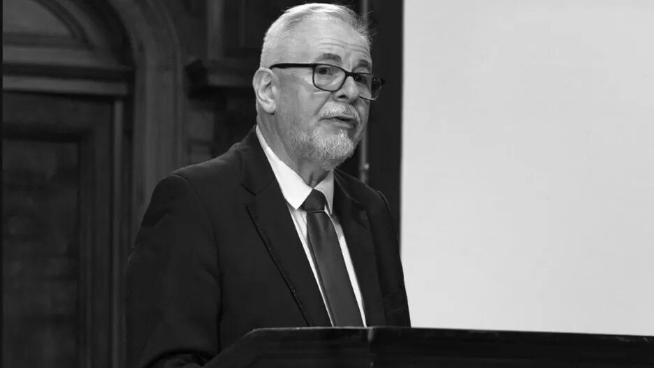 Na zdjęciu widać starszego mężczyznę w garniturze, białej koszuli i krawacie, stojącego za mównicą. Ma krótkie siwe włosy, brodę i okulary, a jego twarz wyraża skupienie podczas przemówienia.