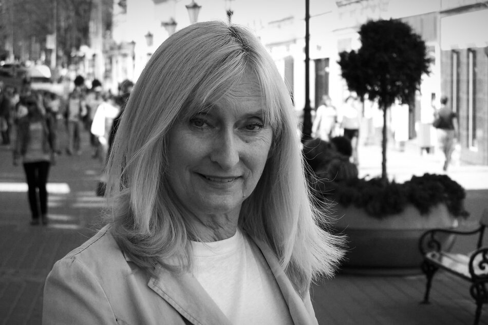 Na zdjęciu widać kobietę w średnim wieku z jasnymi, prostymi włosami, uśmiechającą się lekko do obiektywu. Stoi na ulicy pełnej przechodniów, w słoneczny dzień, ubrana w jasny płaszcz i białą bluzkę.