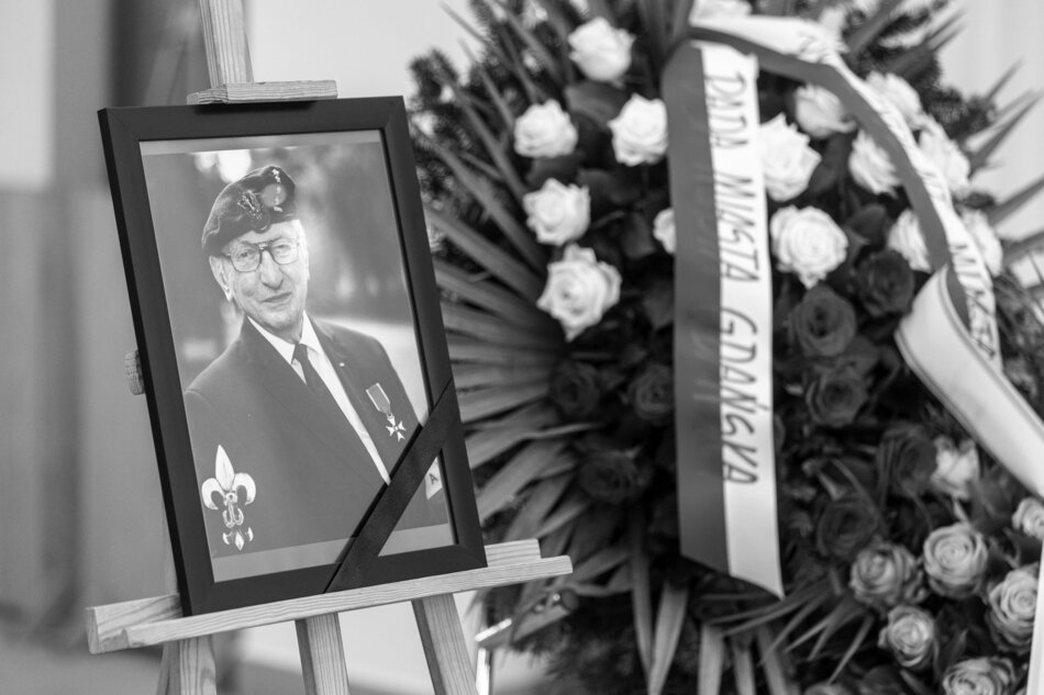 Na zdjęciu widoczna jest portretowa fotografia starszego mężczyzny w mundurze harcerskim z beretem i orderem, ustawiona na sztaludze i przewiązana czarną wstążką żałobną. Obok znajduje się wieniec z biało-czerwonymi kwiatami oraz szarfą z napisem Rada Miasta Gdańska, świadczący o uroczystości pogrzebowej. 