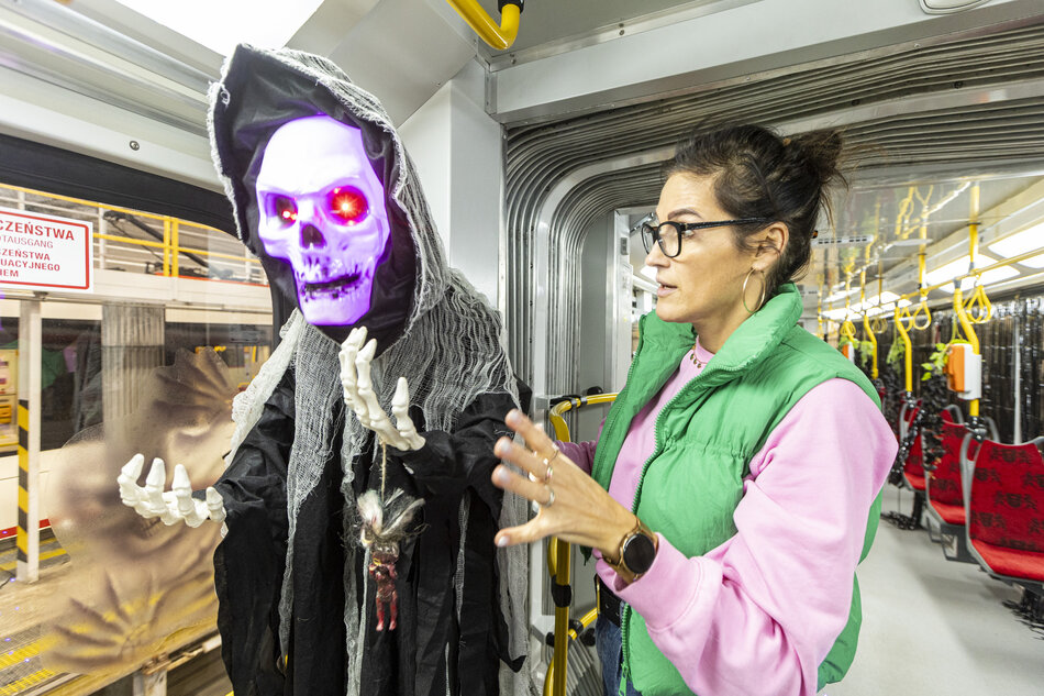 Kobieta w zielonej kamizelce i różowej bluzie stoi obok figury przypominającej upiornego kostuchę z podświetloną, fioletową czaszką i świecącymi oczami. Wnętrze pojazdu udekorowane jest elementami nawiązującymi do Halloween, co tworzy mroczny i zabawny klimat. 