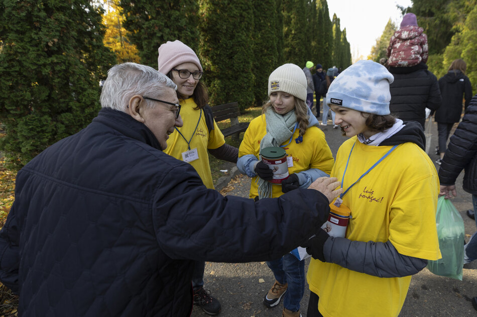 W jesiennym otoczeniu starszy mężczyzna wspiera kwestę, wrzucając datek do puszki trzymanej przez wolontariuszki w żółtych koszulkach. Uśmiechy i serdeczna atmosfera podkreślają zaangażowanie młodych ludzi w akcję charytatywną. 