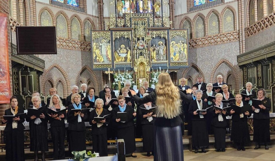 Chór seniorów śpiewa w gotyckim kościele przed bogato zdobionym ołtarzem, prowadzony przez dyrygentkę z długimi blond włosami. Występ odbywa się w podniosłej atmosferze, w otoczeniu witraży i złoconych elementów architektury sakralnej. 