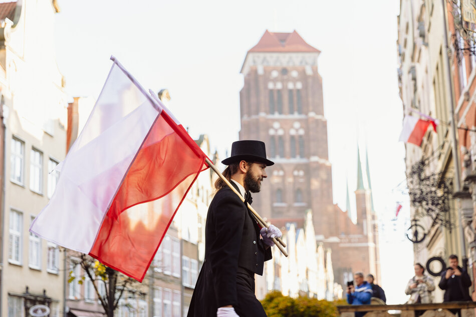 Mężczyzna ubrany w elegancki czarny frak i cylinder niesie biało-czerwoną flagę podczas obchodów Święta Niepodległości w Gdańsku. W tle widać ulicę Długą z charakterystyczną wieżą Bazyliki Mariackiej i obserwujących uczestników. 