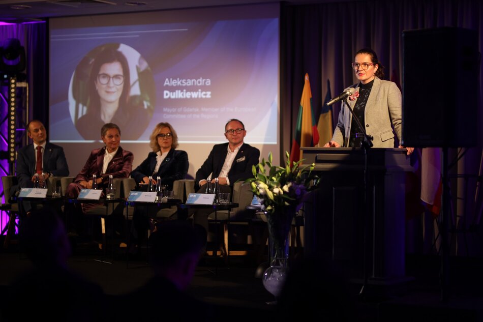 Na zdjęciu widać uczestników panelu dyskusyjnego, a przy mównicy przemawia kobieta w szarej marynarce. W tle znajduje się ekran z jej wizerunkiem i imieniem, a obok na scenie siedzi kilku prelegentów w eleganckich strojach. 