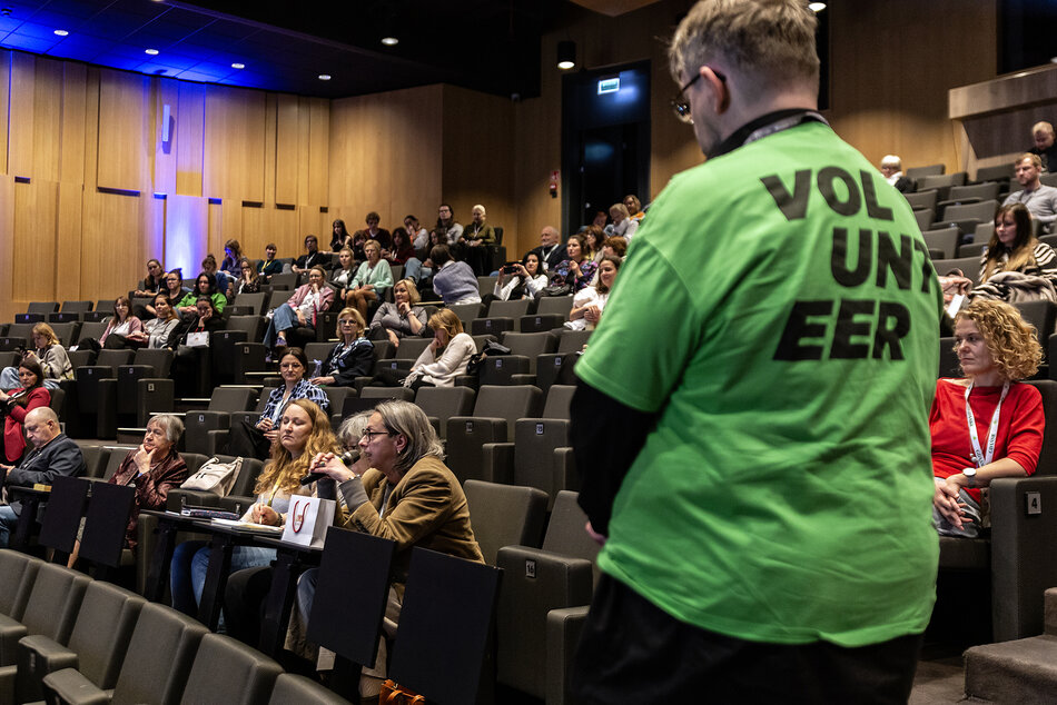 Na sali audiencyjnej siedzi grupa osób, jedna kobieta mówi do mikrofonu, na pierwszym planie stoi mężczyzna w zielonej koszulce z napisem na plecach volunteer.