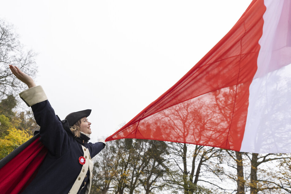 Osoba w stroju stylizowanym na historyczny z uniesionymi rękami trzyma dużą biało-czerwoną flagę rozwianą na wietrze. W tle widać jesienne drzewa i jasne niebo, co podkreśla uroczysty charakter chwili. 