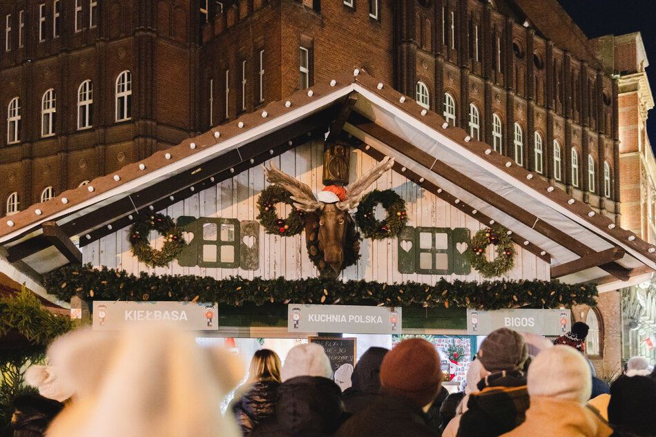 Tłum ludzi gromadzi się przed świątecznym stoiskiem z tradycyjną kuchnią polską, ozdobionym girlandami i wieńcami bożonarodzeniowymi. Nad wejściem wisi głowa łosia w czerwonej czapce Mikołaja, dodając stoisku humorystycznego, zimowego charakteru. 