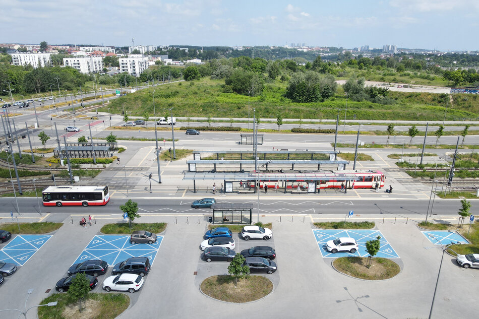 zdjęcie z drona, w dolnej części widać parking z samochodami, jest kilka miejsc wymalowanych na niebisko dla niepełnosprawnych, dalej widać wiaty przystankowe i stojące przy nich dwa biało-czerwone autobusy, w tle zielony teren