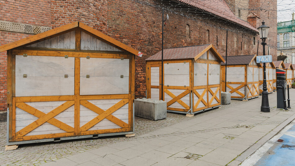 drewniane, zadaszone kramy ustawione na brukowanej ulicy, na tle starego ceglanego muru. Kramy są puste, nad nimi widoczne, nie działające girlandy z ozdobnych lampek