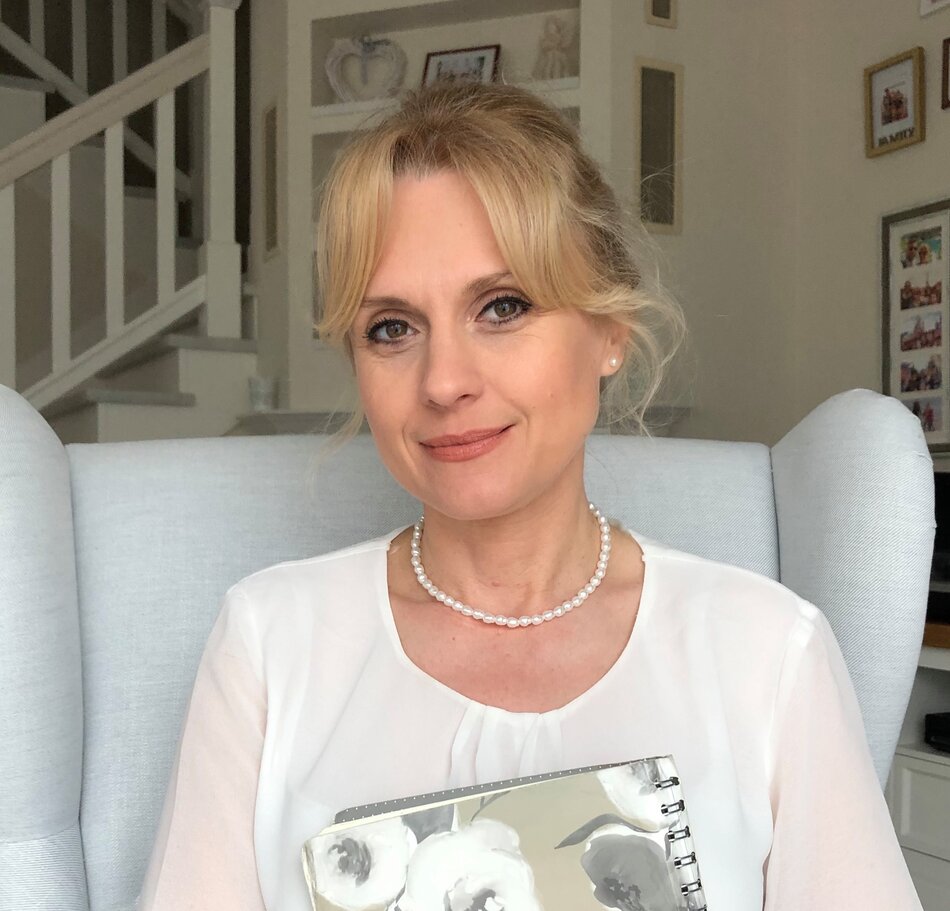 kobieta siedząca na jasnym fotelu, ubrana w elegancką bluzkę, trzymająca notes w kwiatowy wzór; w tle wnętrze z dekoracjami i schodami.