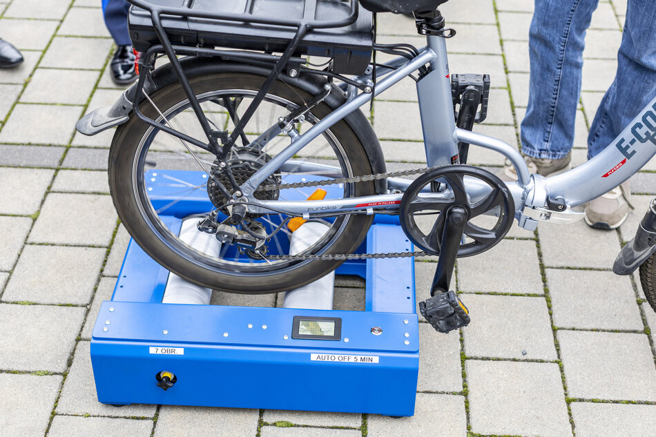 na zdjęciu koło roweru ustawione na urządzeniu w kolorze niebieskim, leży ono na chodniku