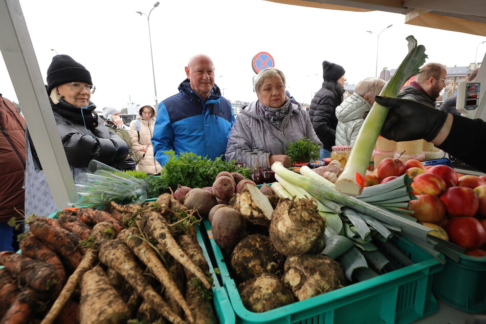 na zdjęciu stragan ze skrzyniami wypełnionymi warzywami, widać pietruszkę, marchew a także zielonego pora, którego ktoś trzyma w ręce, przed straganem stoją trzy starsze osoby