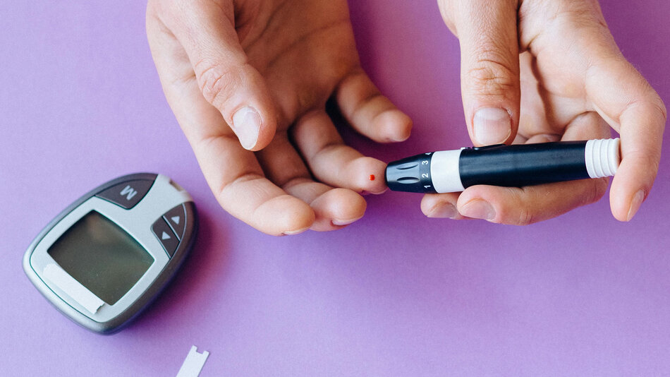 ręka w trakcie badania na poziom cukru. Na zdjęciu widać ukucie i małą plamkę krwi na palcu. obok urządzenie elektroniczne z ekranem