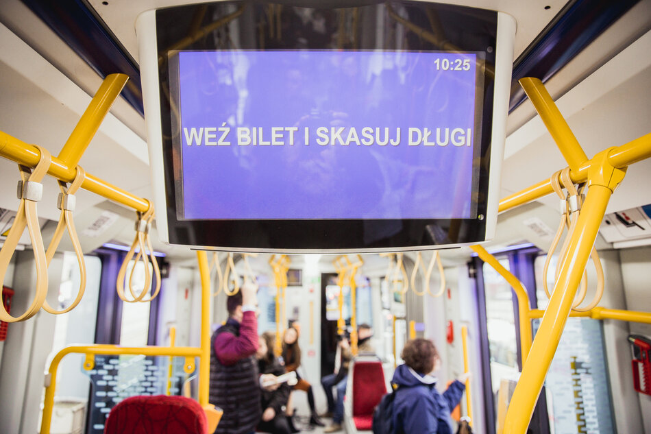 Wnętrze tramwaju, na pierwszym planie ekran z napisem Weź bilet i skasuj długi.