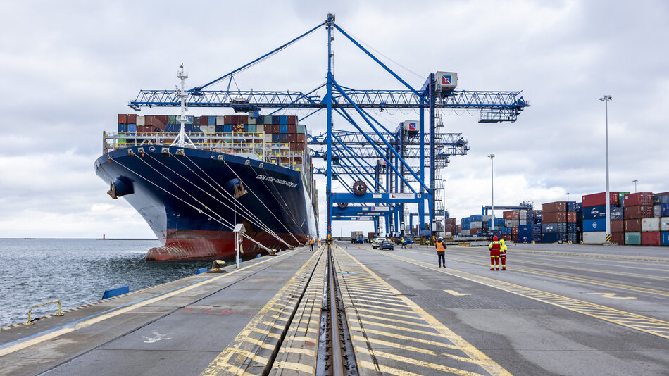 Duży statek zacumowany przy betonowym nabrzeżu, nad nim pracują suwnice, które wyładowują kontenery. Na pierwszym planie widoczne tory