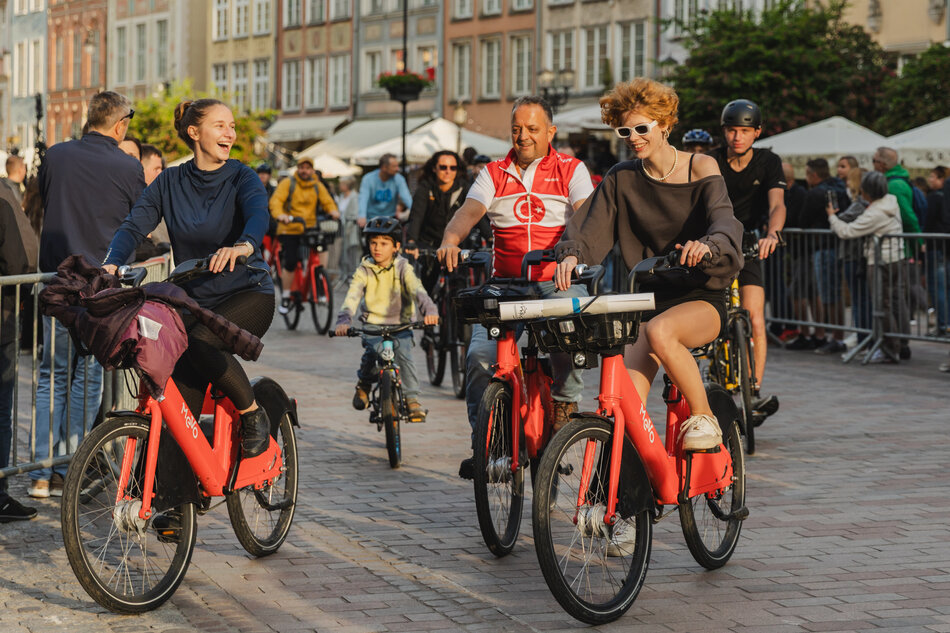 Na zdjęciu widać grupę uśmiechniętych osób jadących na czerwonych rowerach miejskich Mevo ulicą w centrum miasta, w otoczeniu zabytkowych kamienic i licznych przechodniów. Atmosfera jest radosna i pełna energii, a uczestnicy sprawiają wrażenie, że wspólna przejażdżka sprawia im dużo przyjemności. 
