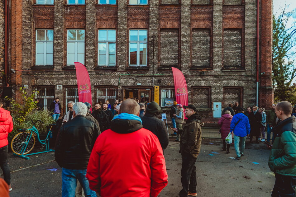 Grupa osób zgromadzona na dziedzińcu przed starym ceglanym budynkiem; ludzie stoją w kolejkach i rozmawiają, obok widoczne dwie czerwone flagi z napisem Gdańsk oraz zaparkowany rower