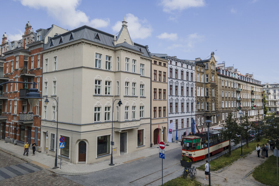  Scena uliczna w europejskim mieście. Na ulicy znajduje się kilka budynków, w tym duży biały budynek z czarnym dachem i wieżą zegarową na górze. Budynki są różnych rozmiarów i kolorów, z których niektóre są czerwone, białe i beżowe. Po prawej stronie jest czerwono-biały wózek na brukowanej ulicy. Ludzie mogą spacerować po chodniku i chodzić. W tle niebo jest niebieskie, widoczne są drzewa i inne budynki.