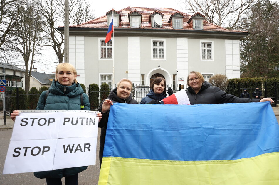 na zdjęciu cztery kobiety protestują pod jasnym budynkiem konsulatu, po prawej dwie kobiety trzymają żółto niebieską flagę, po lewej trzymają białą kartę z czarnym napisem