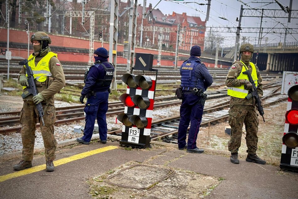 Kilka osób w mundurach i z karabinami oraz zamaskowanymi twarzami stoi na peronie olejowym, w tle tory kolejowe.