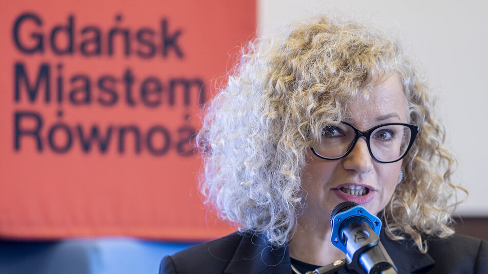  kobieta o kręconych blond włosach i okularach mówiąca do mikrofonu. Ma na sobie czarną bluzę i wydaje się wygłaszać przemówienie lub prezentację. Tło jest zamazane, ale wydaje mi się, że stoi przed czerwonym banerem z białym tekstem Gdańsk Miastem Rowność.
