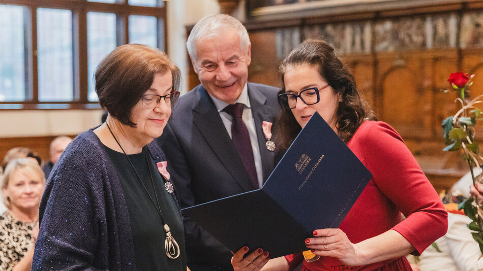  Trzy osoby, dwie kobiety i starszy mężczyzna, stojący w dużym pokoju z wysokim sufitem i ozdobnymi dekoracjami. Kobieta po lewej nosi niebieski sweter i naszyjnik, a kobieta pośrodku trzyma czarny folder z pieczęcią to młoda kobietka w pomarańczowej sukience i okularach, która również trzyma folder. Oboje uśmiechają się i wydają się gratulować mężczyźnie jego osiągnięć. W tle widzimy grupę ludzi siedzących na krzesłach, z których niektórzy trzymają płatki róży.