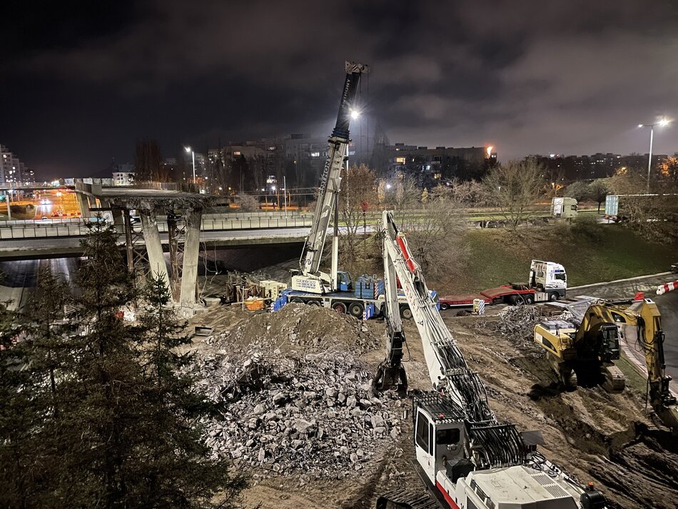 Na zdjęciu widać nocne prace rozbiórkowe przy wiadukcie, z udziałem ciężkiego sprzętu budowlanego – koparek, dźwigów i maszyn kruszących beton. Konstrukcja mostu jest częściowo zdemontowana, a wokół leżą sterty gruzu i odłamków betonu, oświetlone silnymi lampami roboczymi. 