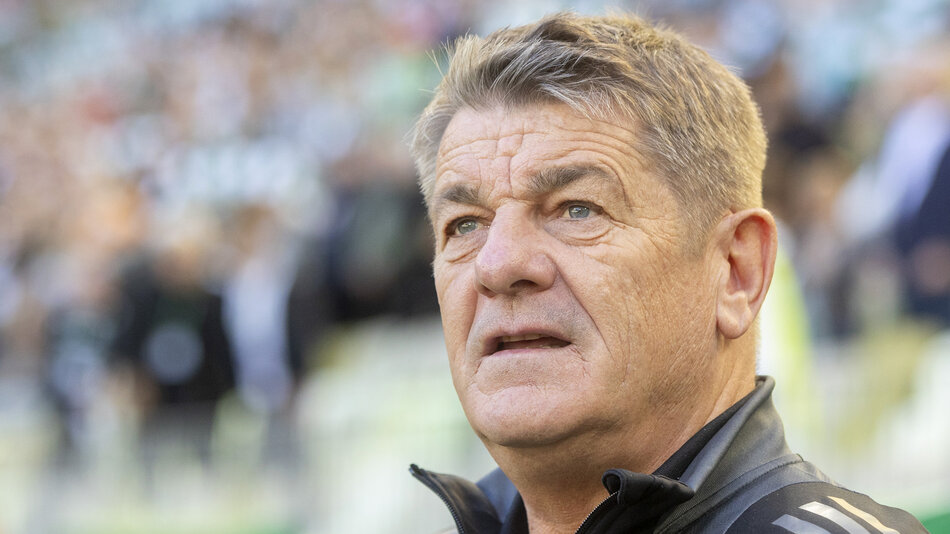  mężczyzna w średnim wieku stoi na boisku piłkarskim, nosi czarną kurtkę z logo, patrzy na bok z poważnym wyrazem twarzy, w tle są niewyraźne figury widzów na trybunach.