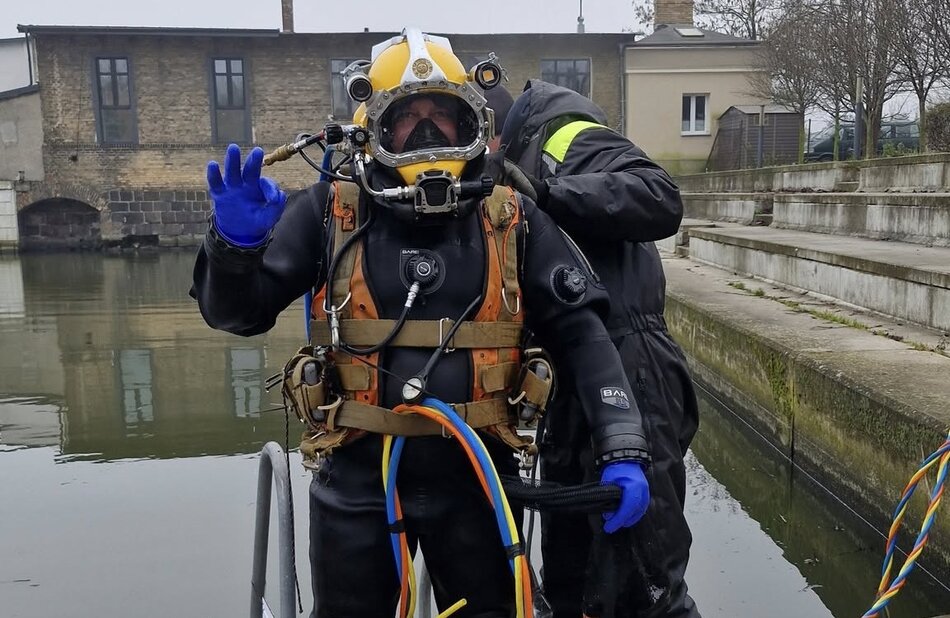 Osoba w profesjonalnym skafandrze nurkowym stoi na pomoście, unosząc rękę w geście sygnału OK. Druga osoba pomaga jej z wyposażeniem, a w tle widać spokojny akwen i zabudowania portowe. 