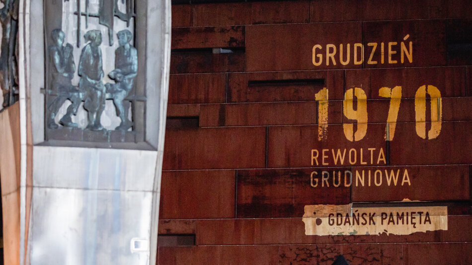  zbliżenie ściany z napisem GRUDZIEN 1970 Rewolta Grudniowa napisanym w złocie. Tekst jest w języku polskim i tłumaczy się na Gdańską Pamietę w jęz. angielskim. Po lewej stronie znajduje się metalowa tablica z grupą osób stojących na piedestale, która wydaje się być pomnikiem lub pomnikiem osiemdziesiątych lub osiedemdziesiątego wieku w Polsce.