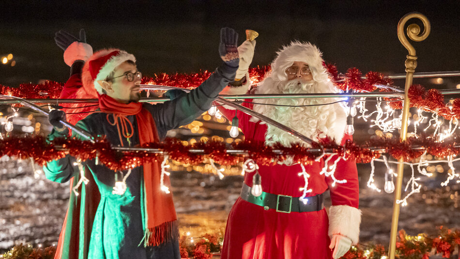  Dwie osoby ubrane jak Święty Mikołaj i Pani Mikołaj na łodzi w nocy.Osoba po lewej nosi czerwono-zielony strój z kapeluszem i szalikiem Świętego Mikołaja i trzyma dzwonek w ręku. Ma również okulary i duży uśmiech na twarzy.Po prawej stronie jest osoba ubrana jak ta sama osoba, ubrana w tradycyjny czerwony garnitur z białym futrem i czarnym pasem. Stoją za szynką pokrytą czerwoną tęczą i girlandą, z wiszącymi z niej światłami bożonarodzeniowymi.W tle możemy zobaczyć śnieżny krajobraz z drzewami i budynkami oświetlonymi kolorowymi światłami.