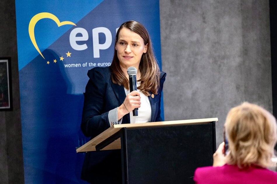 Przemawia kobieta w średnim wieku - ma długie ciemne włosy