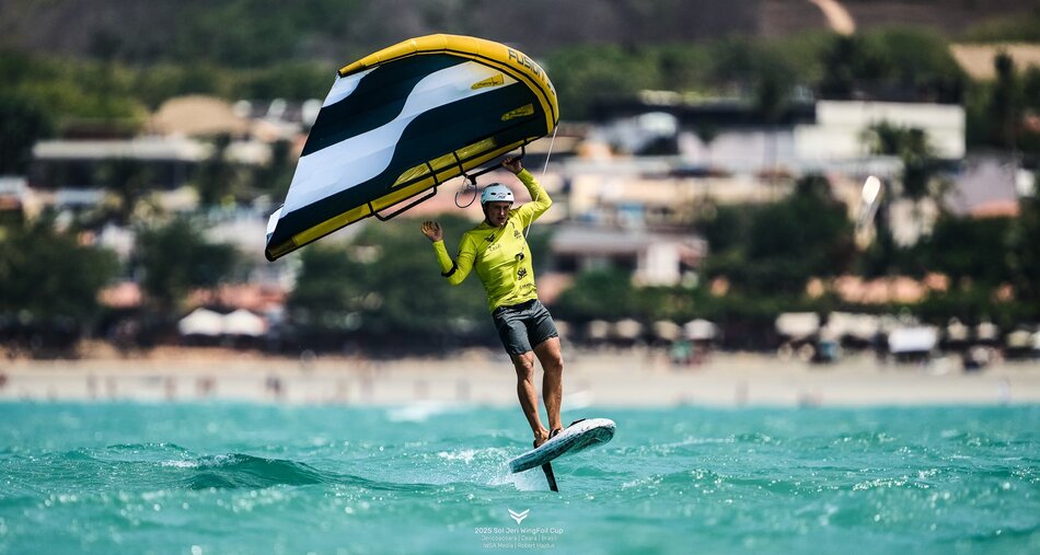 Zawodnik w żółtej koszulce leci nad wodą na desce wingfoil, trzymając skrzydło uniesione nad głową. W tle widać zabudowania nadmorskiego miasteczka, a jasna woda podkreśla efekt lekkości lotu nad falami.