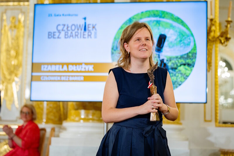 Kobieta w eleganckiej, granatowej sukni stoi w reprezentacyjnej sali i trzyma w dłoniach statuetkę wręczoną podczas uroczystości. W tle widoczny jest ekran z grafiką wydarzenia oraz fragmenty złoconych dekoracji wnętrza. 