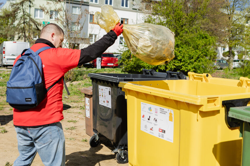 na zdjęciu mężczyzna w czerwonej kurtce i niebieskim plecaku wrzuca żółtą plastikową torbę do żółtego kosza na śmieci