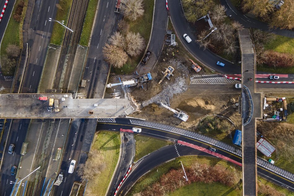 Zdjęcie wykonane z lotu ptaka ukazuje rozbiórkę fragmentu kładki lub wiaduktu nad rozbudowanym węzłem drogowym, z widocznym gruzowiskiem i pracującymi maszynami budowlanymi. Ruch samochodowy odbywa się po częściowo przeorganizowanych jezdniach, co wskazuje na prowadzenie robót przy zachowaniu przejezdności głównych tras. 