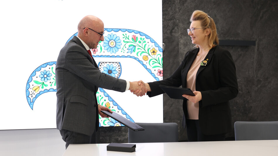 Mężczyzna w szarym garniturze i kobieta w czarnym żakiecie podają sobie dłonie, patrząc na siebie z uśmiechem, jak podczas oficjalnego podpisania umowy. Oboje trzymają czarne teczki, a za nimi widać jasne tło z kolorowym, kwiatowym wzorem.