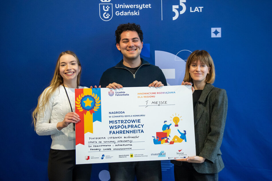 Dwie młode kobiety i jeden młody mężczyzna pozują do zdjęcia, uśmiechając się. Trzymają w rękach tablicę informującą o zwycięstwie w konkursie naukowym.