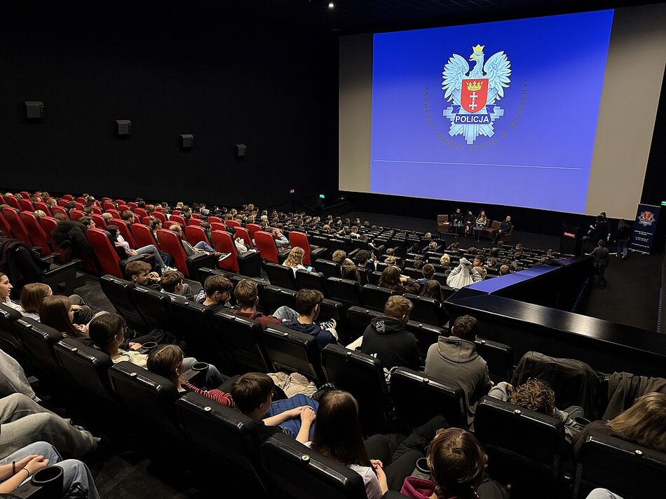 Sala kinowa. Na fotelach siedzą ludzie. Na ekranie wyświetla się logo policji.