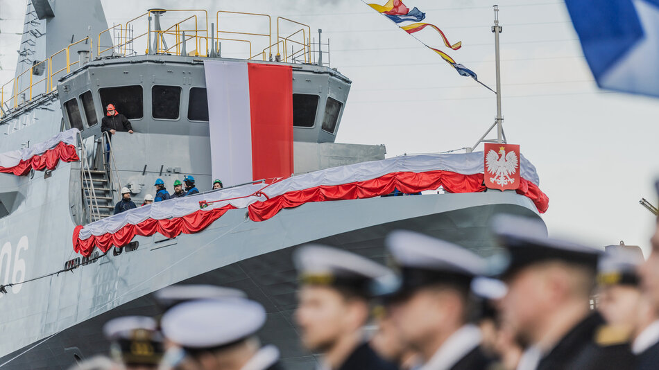  Duży statek z czerwoną i białą flagą zasłonioną na boku; statek wydaje się być statkiem marynarki wojennej, ponieważ na pokładzie jest kilka osób; jest to grupa mężczyzn w mundurach wojskowych, wszyscy w białych czapki i mundury z czerwonymi odznakami; stoją w linii, skierowani w tym samym kierunku i wydają się oglądać odbywającą się paradę lub ceremonię.