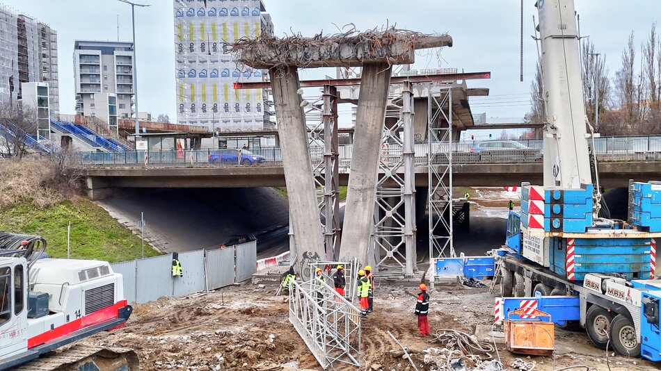 Na zdjęciu widoczne są prace rozbiórkowe fragmentu estakady, z odsłoniętymi żelbetowymi podporami i pozostałościami zbrojenia na górnej krawędzi konstrukcji. Wokół miejsca robót znajdują się pracownicy w odzieży ochronnej oraz ciężki sprzęt budowlany, a w tle widoczna jest czynna infrastruktura drogowa i zabudowa mieszkaniowa. 