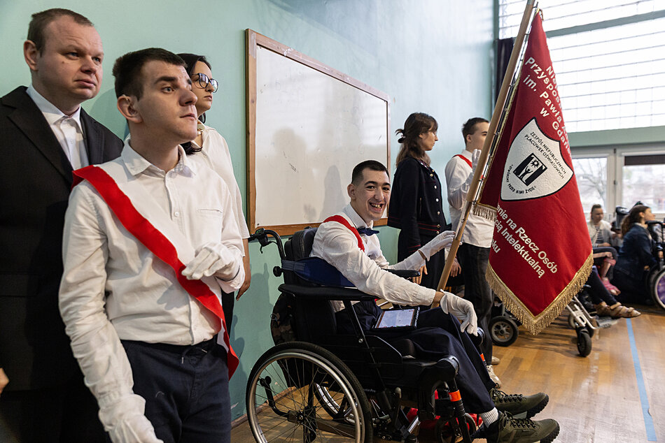 Poczet sztandarowy na uroczystości szkolnej na sali gimnastycznej. sztandar trzyma uczeń na wózku.