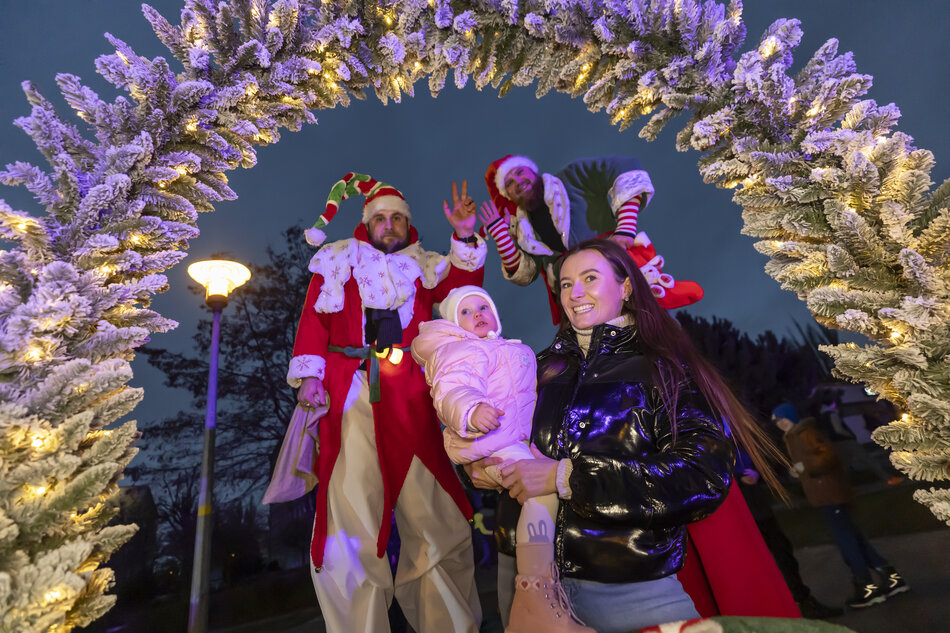 Kobieta z dzieckiem na rękach pozuje pod podświetloną, świąteczną bramą udekorowaną sztucznym śniegiem i lampkami. W tle widać animatorów w kostiumach świątecznych elfów oraz uczestników wydarzenia zgromadzonych na świeżym powietrzu. 