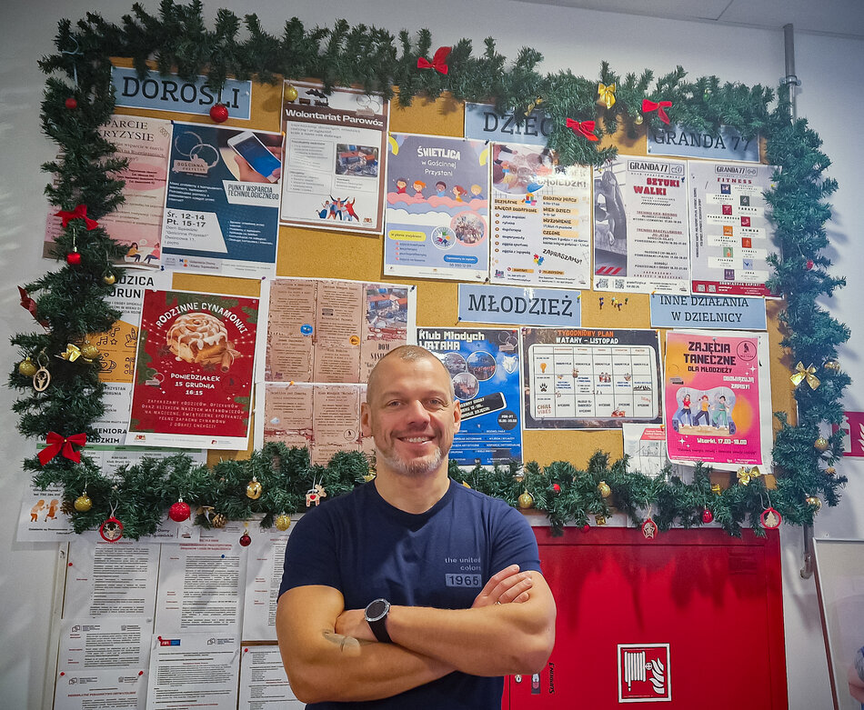 Mężczyzna około czterdziestki, za nim na tablicy plakaty z zajęciami podzielonymi wiekowo: dla dzieci, młodzieży, dorosłych i seniorów