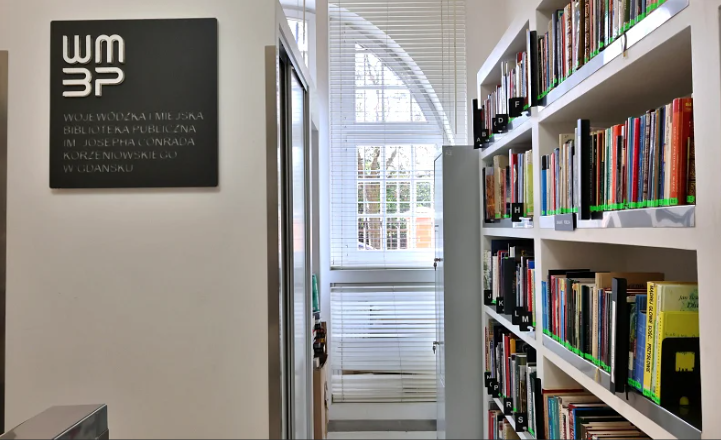 Wnętrze Wojewódzkiej i Miejskiej Biblioteki Publicznej w Gdańsku. Na białej ścianie widać czarną tablicę z logotypem WiMBP, a obok białe regały wypełnione książkami. W tle znajduje się wysokie okno z białymi żaluzjami.